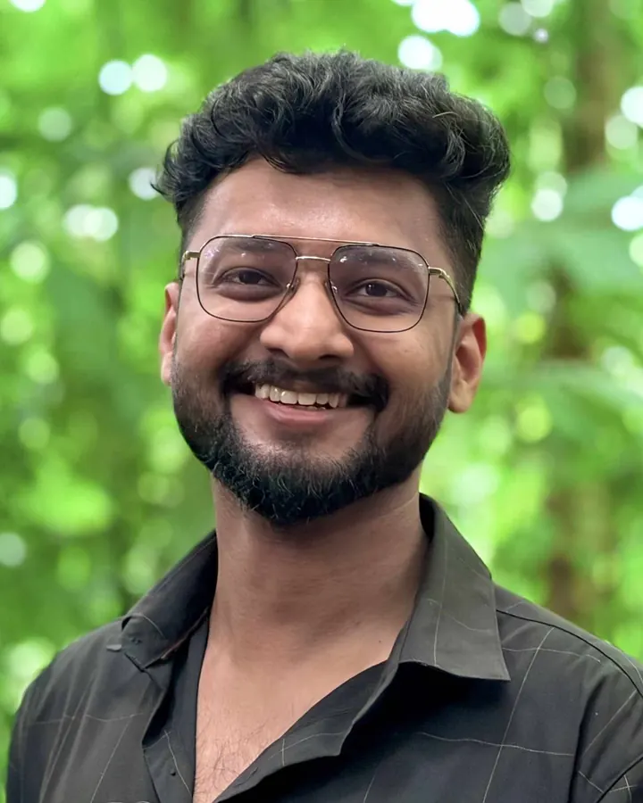 Arun Prakash, smiling, in glasses and a dark shirt, with green leaves behind him
